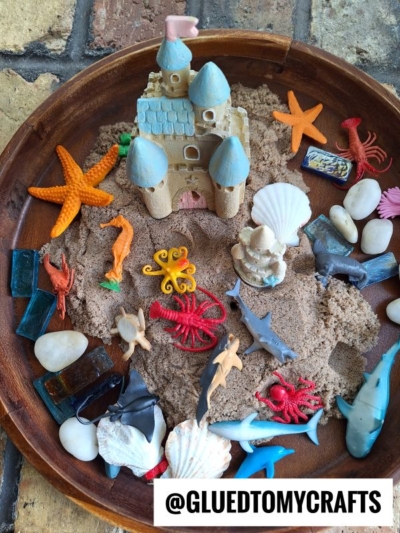 A round wooden tray filled with sand, a toy sandcastle, plastic sea animals, seashells, starfish, rocks, and blue glass pieces—perfect for inspiring kinetic sand play ideas. @GLUEDTOMYCRAFTS text at the bottom.