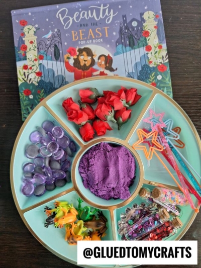A round teal tray holds purple gems, red artificial roses, star wands, animal figurines, craft beads, jars with glitter, and purple kinetic sand—perfect for creative kinetic sand play ideas. Behind it is a Beauty and the Beast pop-up book.