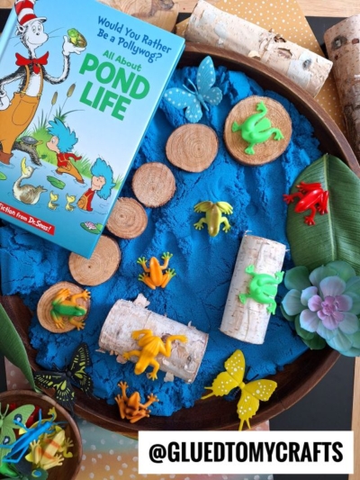 A colorful sensory tray features blue kinetic sand, toy frogs, butterflies, wood slices, artificial leaves, and the Dr. Seuss book “All About Pond Life,” offering playful pond-themed kinetic sand play ideas.