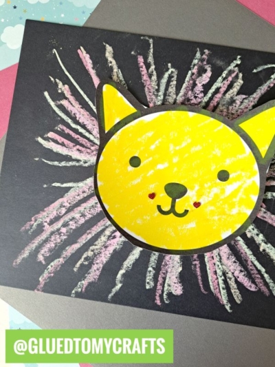 A child’s craft of a yellow cat face made with crayons on black paper, surrounded by colorful chalk art projects lines resembling a mane. The Instagram handle @GLUEDTOMYCRAFTS appears at the bottom.