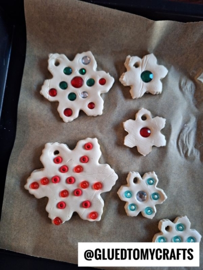 Five unbaked salt dough ornaments shaped like snowflakes, decorated with red, green, and clear beads, are arranged on a parchment-lined baking tray. Text @GLUEDTOMYCRAFTS is visible in the bottom right corner.