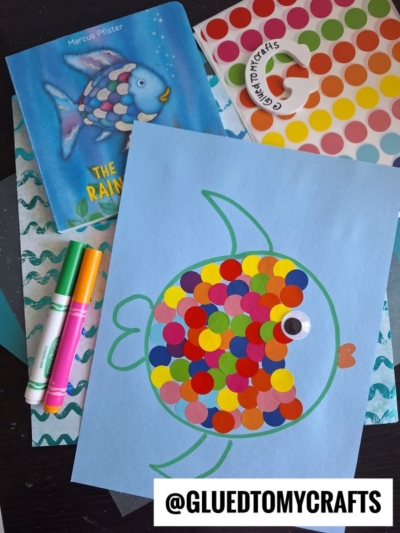 A children’s art project inspired by The Rainbow Fish book shows a paper fish made with colorful dot stickers, a googly eye, and coding label crafts on blue paper. Markers, the book, and craft supplies surround the artwork. @GLUEDTOMYCRAFTS is written below.