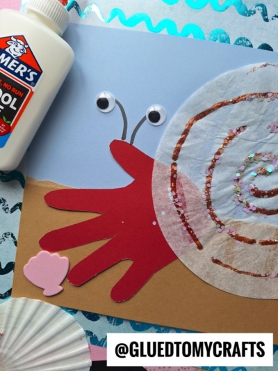 A childrens snail craft featuring a red handprint as the snails body, a googly eye, and a swirled, glittery paper shell, with white glue, cupcake liners, and the @gluedtomycrafts handle visible.