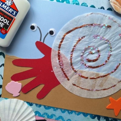 A handprint snail craft made with a red paper hand, brown body, coffee filter shell with glitter, googly eyes, glue bottle, seashells, and ocean-themed background. Text @GLUEDTOMYCRAFTS is at the bottom.