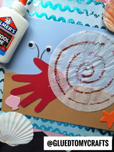 A handprint snail craft made with a red paper hand, brown body, coffee filter shell with glitter, googly eyes, glue bottle, seashells, and ocean-themed background. Text @GLUEDTOMYCRAFTS is at the bottom.