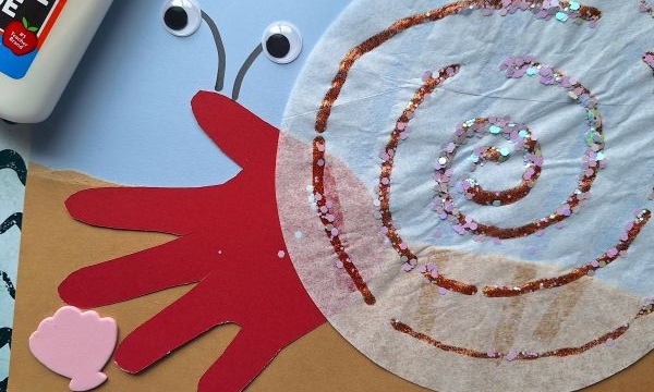 A handprint snail craft made with a red paper hand, brown body, coffee filter shell with glitter, googly eyes, glue bottle, seashells, and ocean-themed background. Text @GLUEDTOMYCRAFTS is at the bottom.