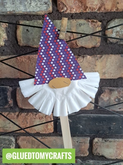 A handmade gnome craft, perfect for coffee filter crafts for kids, features a star-patterned paper hat, coffee filter beard, and oval nose on a stick—clipped to wire against a brick wall with a green @GLUEDTOMYCRAFTS label at the bottom.