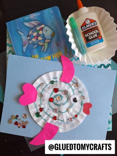 A Rainbow Fish book, glue, sequins, coffee filter crafts for kids, and a paper plate fish with pink fins, blue swirls, and gem decorations are arranged on a table. @GLUEDTOMYCRAFTS is labeled at the bottom.