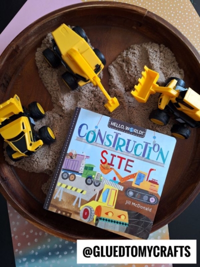 A round wooden tray filled with kinetic sand, three yellow toy construction vehicles, and a children’s book titled Construction Site by Jill McDonald—perfect for inspiring new kinetic sand play ideas. @GLUEDTOMYCRAFTS is displayed at the bottom.