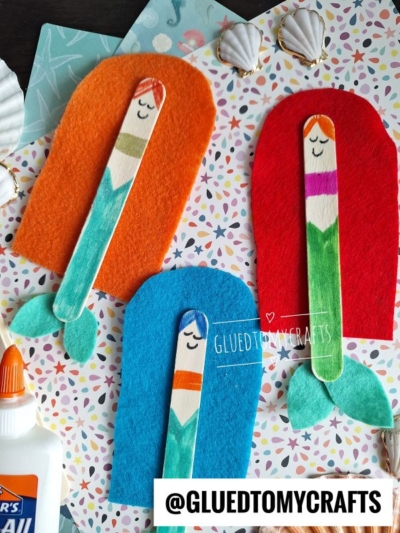 Three handmade mermaid crafts use colored felt for tails and decorated popsicle sticks for bodies, displayed on patterned paper. A bottle of glue and seashells are nearby, with @GLUEDTOMYCRAFTS text at the bottom.