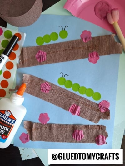 A HUNGRY CATERPILLAR CRAFT project with brown crepe paper strips, green sticker caterpillars, pink paper spots, a glue bottle, paint, sponge dauber, and colored paper neatly arranged on a table. @GLUEDTOMYCRAFTS is labeled at the bottom.