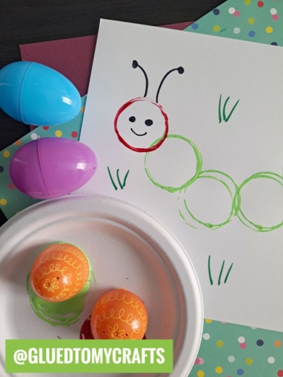 A fun Plastic Egg Stamp Crafts project: create a caterpillar by stamping green circles with plastic Easter eggs, adding a red head and smiley face. Colorful and decorated orange eggs surround the scene, labeled @GLUEDTOMYCRAFTS.
