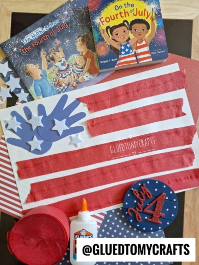 A handmade American flag craft with red tissue paper stripes, white paper stars, and a blue handprint is surrounded by 4th of July crafts, themed children's books, colorful craft supplies, and patriotic decorations.