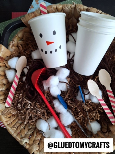 A basket with two white paper cups (one decorated as a snowman), cotton balls, wooden spoons with red stripes, brown yarn, craft tools, and shredded paper—perfect for winter sensory bins. The handle @GLUEDTOMYCRAFTS is at the bottom.