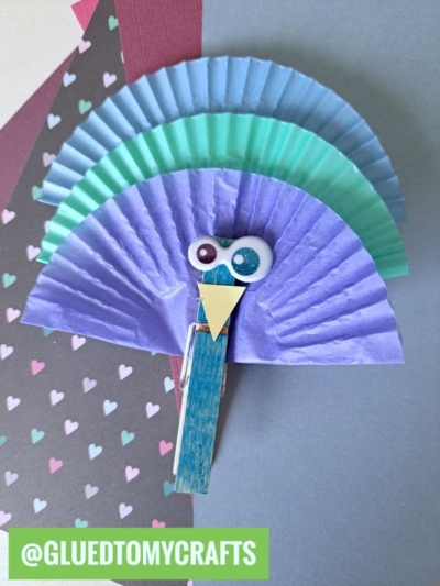A colorful peacock craft made with layered blue and purple cupcake liners, googly eyes, and a clothespin for the body—perfect for clothespin crafts for kids—displayed on a pastel background with a tag that reads @GLUEDTOMYCRAFTS.