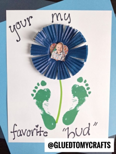 A handmade HANDPRINT FATHER'S DAY card featuring blue cupcake liners as flower petals, a child’s photo in the center, green painted footprints as leaves, and “You’re my favorite bud” written in black marker.