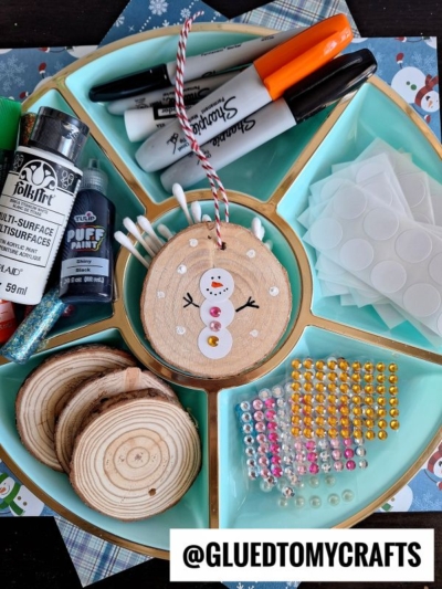 A divided tray holds craft supplies: wood slices, markers, paint, glue, cotton swabs, sticker gems, and foam dots. In the center is a snowman wooden ornament with buttons and sequins. A coding label crafts tag @GLUEDTOMYCRAFTS sits at the bottom.