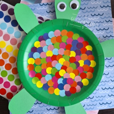 A craft turtle made from a green paper plate decorated with colorful dot stickers, green paper head, tail, and legs. Coding label crafts supplies and sheets of stickers are on the table beside it.