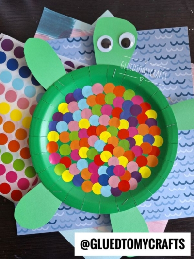 A craft turtle made from a green paper plate decorated with colorful dot stickers, green paper head, tail, and legs. Coding label crafts supplies and sheets of stickers are on the table beside it.