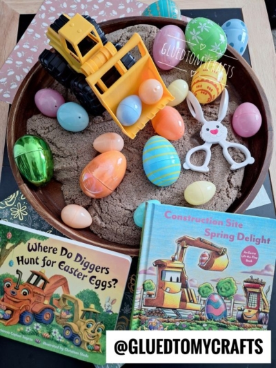 A bowl filled with kinetic sand, plastic Easter eggs, a toy dump truck, and bunny scissors offers fun kinetic sand play ideas. Two children’s books—Where Do Diggers Hunt for Easter Eggs? and Construction Site Spring Delight—are nearby.