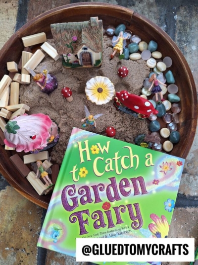 A round tray displays sand, pebbles, wooden blocks, artificial flowers, fairy and mushroom figurines, and a small house—perfect for kinetic sand play ideas. In front is the book How to Catch a Garden Fairy. A tag reads @GLUEDTOMYCRAFTS.