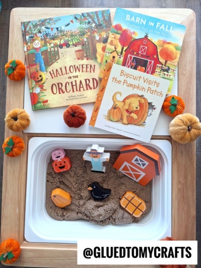 A sensory bin with sand, farm-themed toys, and mini pumpkins sits next to three autumn children’s books: “Halloween in the Orchard,” “Barn in Fall,” and “Biscuit Visits the Pumpkin Patch.”.