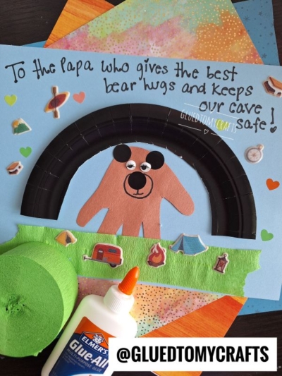 A handmade HANDPRINT FATHER'S DAY card features a bear created from a brown handprint and black paper on a blue background with stickers, hearts, and stars. Text reads, To the Papa who gives the best bear hugs and keeps our cave safe!.