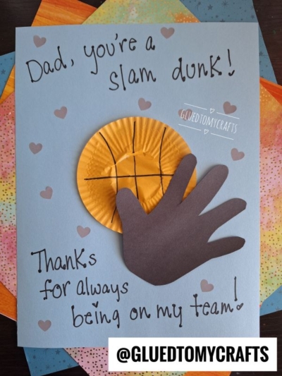A handmade HANDPRINT FATHER'S DAY card features a paper cupcake liner as a basketball and a cutout of a hand. Text reads, Dad, you're a slam dunk! Thanks for always being on my team! The card sits on colorful paper.