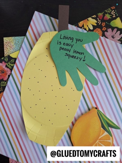 A handmade lemon craft made from yellow and green paper, featuring a green handprint leaf for HANDPRINT FATHER'S DAY and the message Loving you is easy peasy lemon squeezy! on striped and floral backgrounds. @GLUEDTOMYCRAFTS at bottom.
