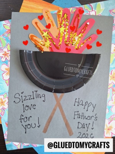 A HANDPRINT FATHER'S DAY card featuring two handprints as a campfire with gold glitter and red hearts. The campfire sits above two brown paper logs and a black paper plate. Sizzling love for you! Happy Fathers Day 2026 is written below.
