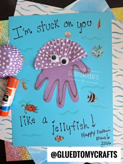 A handmade HANDPRINT FATHER'S DAY card features a purple jellyfish made from a cupcake liner and paper with googly eyes, sea-themed stickers, and the handwritten message: “I’m stuck on you like a jellyfish! Happy Father’s Day!”.