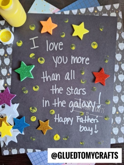 A handmade HANDPRINT FATHER'S DAY card with colorful foam stars and painted yellow dots on black paper. The message reads: I love you more than all the stars in the galaxy! Happy Fathers Day! @GLUEDTOMYCRAFTS at the bottom.