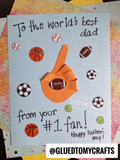 A handmade HANDPRINT FATHER'S DAY card decorated with sports ball stickers and a big orange foam finger. The card says, “To the world’s best dad from your #1 fan! Happy Father’s Day!” @GLUETOMYCRAFTS is printed at the bottom.