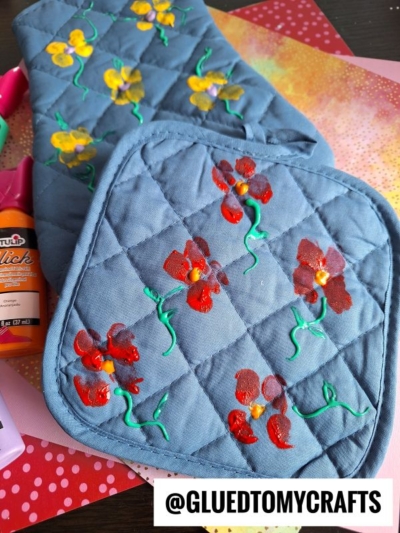 Two quilted blue pot holders are decorated with red and yellow painted flowers and green stems. Craft supplies are visible around them. @GLUEDTOMYCRAFTS is written at the bottom of the image.