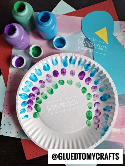 A paper plate with a rainbow made of finger-painted dots in purple, blue, and green, surrounded by paint bottles. Colored paper and a blue paper penguin are in the background. @GLUEDTOMYCRAFTS watermark is visible.