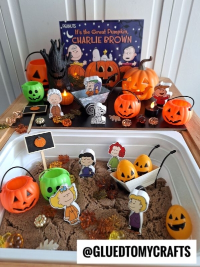 A sensory bin inspired by “It’s the Great Pumpkin, Charlie Brown” featuring kinetic sand play ideas with mini pumpkins, candles, autumn leaves, and Peanuts character cutouts arranged among Halloween decorations.