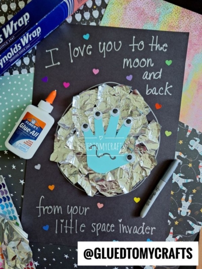 A handmade HANDPRINT FATHER'S DAY card on black paper reads I love you to the moon and back from your little space invader. It features a turquoise handprint with googly eyes, foil, hearts, plus glue and Sharpie nearby.