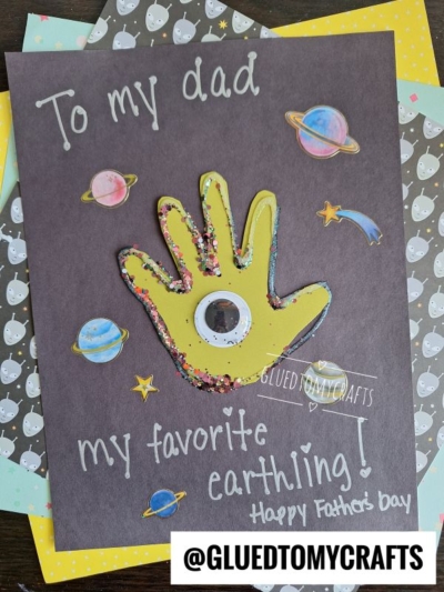A handmade HANDPRINT FATHER'S DAY card reading To my dad, my favorite earthling! Happy Fathers Day, with a yellow handprint, googly eye, and glitter, surrounded by drawings of planets and stars.