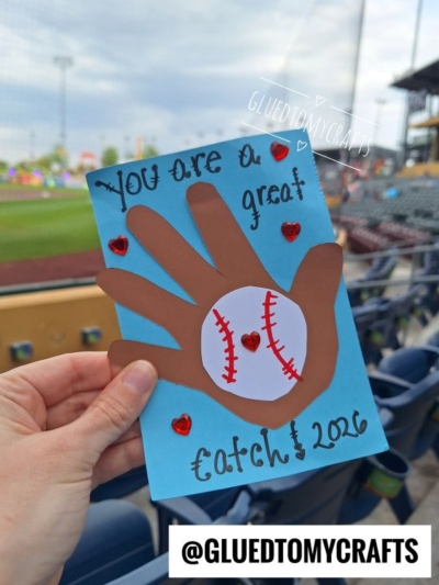 A hand holds a HANDPRINT FATHER'S DAY card with a baseball, red hearts, and the text "You are a great catch! 2026" at a baseball stadium. The handle @GLUETOMYCRAFTS is shown.