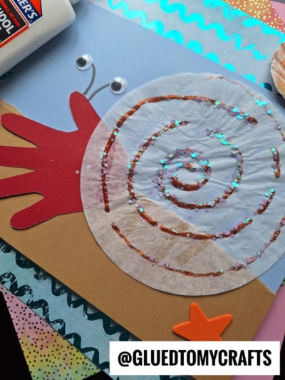 A childrens snail craft made from a coffee filter with glitter, a red handprint as the snail’s body, googly eyes on paper stalks, and colorful paper accents, with a bottle of glue nearby. @GLUEDTOMYCRAFTS is labeled at the bottom.