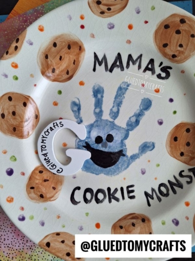 A plate decorated with chocolate chip cookies and the phrase Mamas Cookie Monster. In the center is a blue handprint resembling Cookie Monsters face, with added eyes and a mouth. @GluedToMyCrafts is displayed at the bottom.