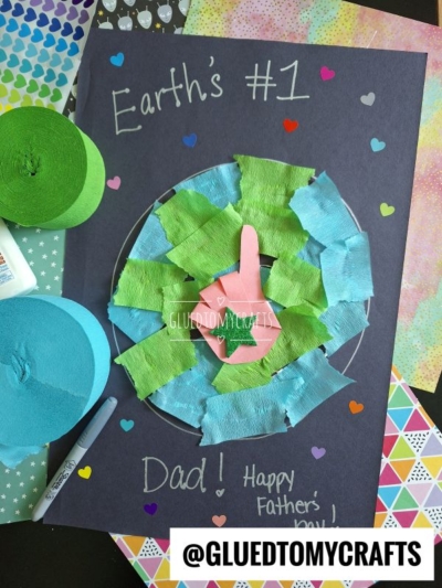 A HANDPRINT FATHER'S DAY craft features a handprint on green and blue tissue paper Earth, with “Earth’s #1 Dad! Happy Father’s Day” in white on dark paper, surrounded by hearts and colorful craft supplies.