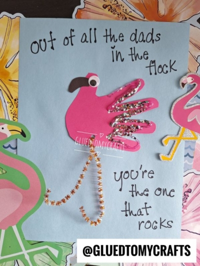 A handmade HANDPRINT FATHER'S DAY card features a pink flamingo crafted from a cut-out handprint and glitter, with a pipe cleaner for legs. The card reads, Out of all the dads in the flock, you're the one that rocks.