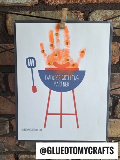 A child's orange handprint is used as flames above a grill drawing with the words Daddy's Grilling Partner, making it a perfect HANDPRINT FATHER'S DAY craft. The image is clipped to a string and displayed on a brick wall. @GLUEDTOMYCRAFTS at the bottom.
