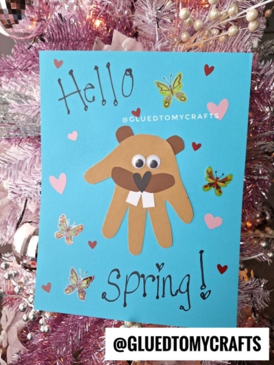 A blue card with a handprint beaver craft, butterfly and heart stickers, and Hello Spring! written on it, is displayed on a pink and white decorated tree. @GLUEDTOMYCRAFTS is printed at the bottom.