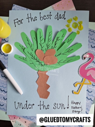 A handmade HANDPRINT FATHER'S DAY card features a palm tree crafted from green and brown paper, a yellow painted sun, coconuts, and the words For the best dad under the sun! Happy Fathers Day! on blue background. A pink flamingo sits beside it.