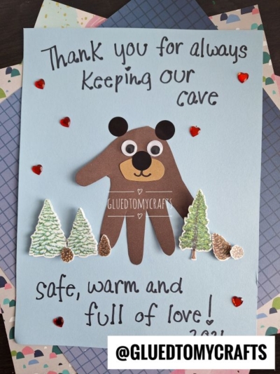 A handmade HANDPRINT FATHER'S DAY card featuring a bear crafted from brown paper, trees, pinecones, red heart stickers, and the message: Thank you for always keeping our cave safe, warm, and full of love! on a blue background.