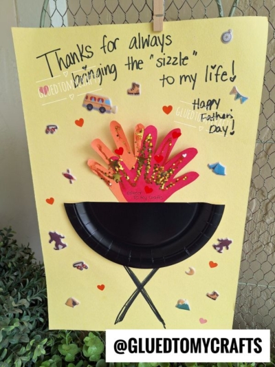 Yellow card with a paper grill and two handprint cutouts as fiery flames, decorated with stickers and hearts. Perfect HANDPRINT FATHER'S DAY craft! Text says “Thanks for always bringing the ‘sizzle’ to my life! Happy Father’s Day!” @GLUEDTOMYCRAFTS.