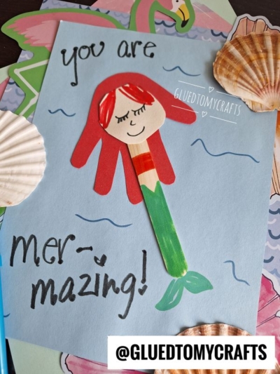 A handmade mermaid craft using a popsicle stick with red paper hair and a drawn face is glued to blue paper with You are mer-mazing! written on it, surrounded by seashells and ocean-themed decorations.