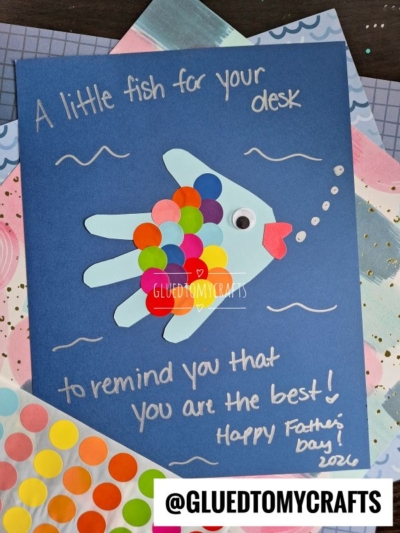 A handmade HANDPRINT FATHER'S DAY card features a colorful fish crafted from paper circles and fins, with the message: “A little fish for your desk to remind you that you are the best! Happy Father’s Day! 2024.” Supplies surround the card.